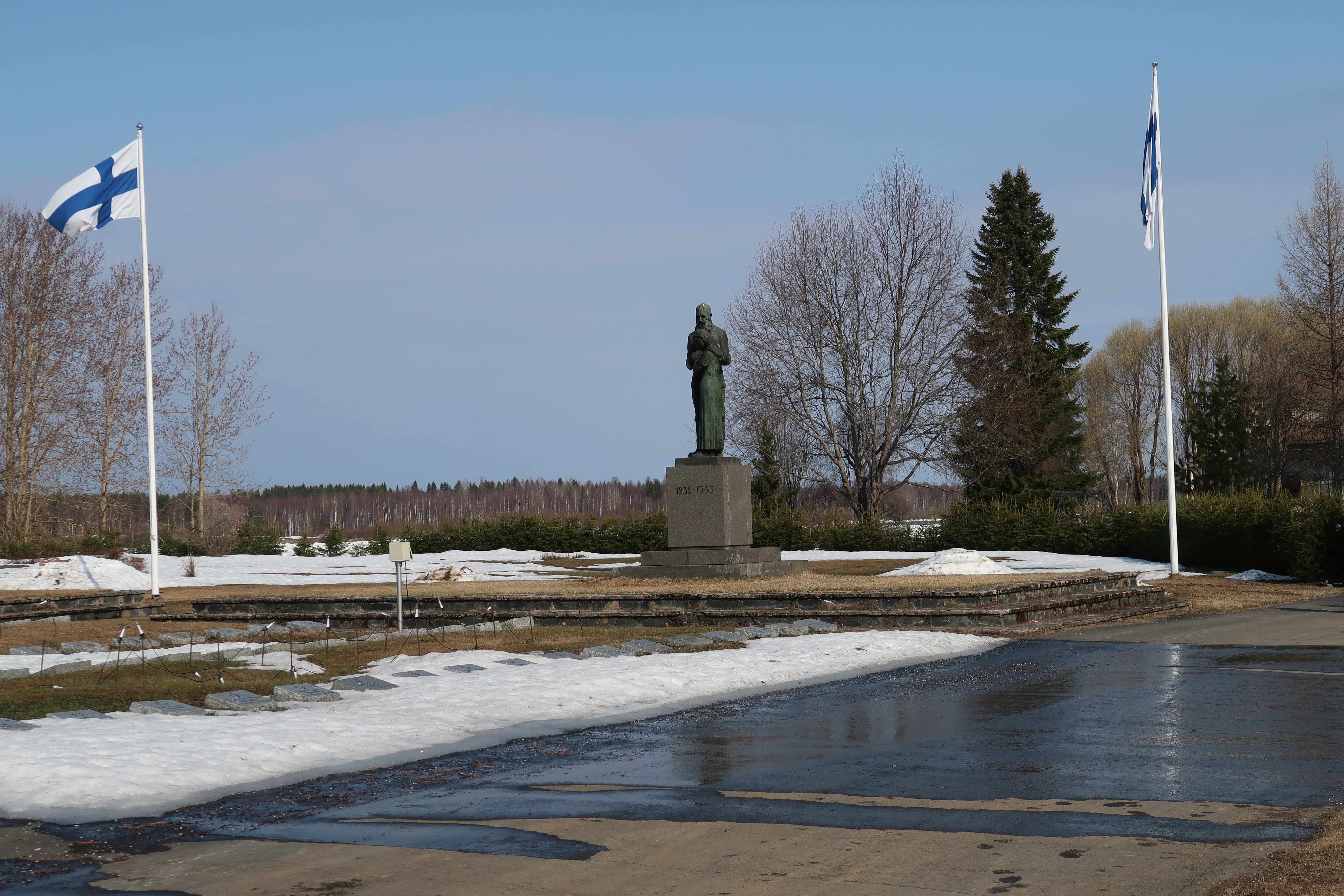 Kunniakäynti sankarihaudalla ilman yleisön läsnäoloa Valokuvassa on Tervolan sankaripatsas ja kaksi liehuvaa Suomen lippua.